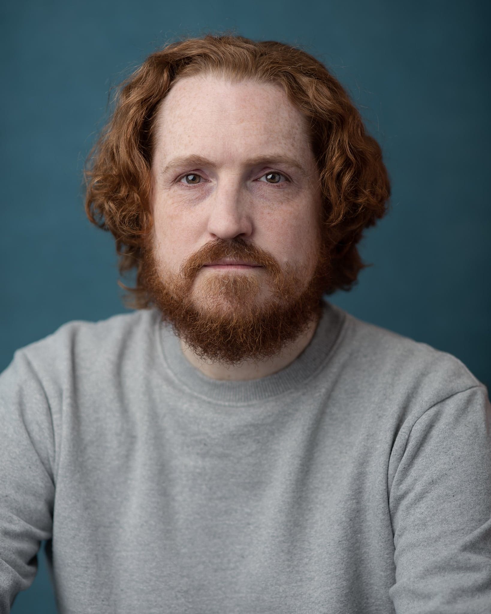 Chest-up headshot of a red-haired, bearded man in a grey sweatshirt against a deep teal hand-painted canvas backdrop